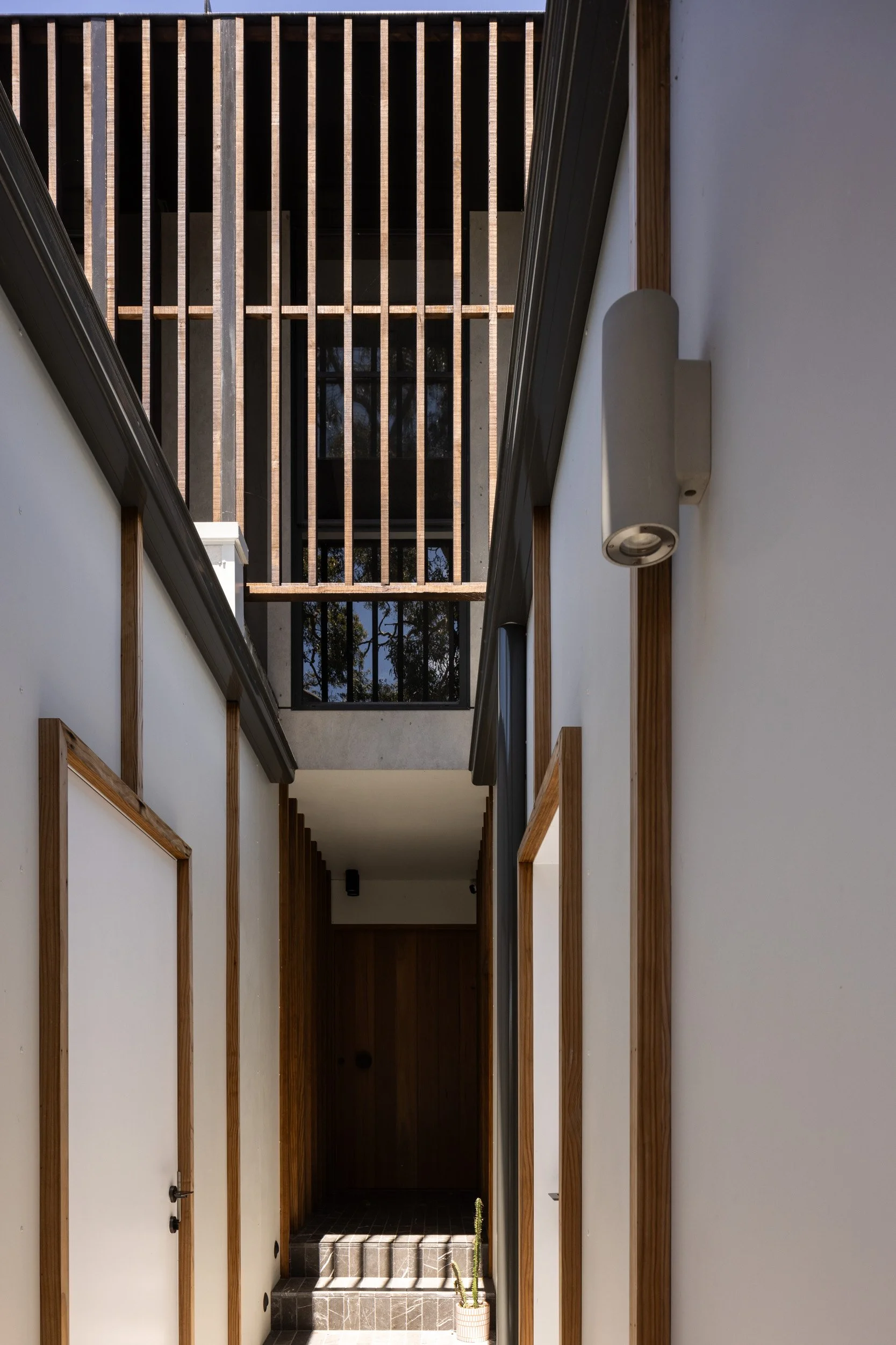 A modern indoor hallway with white walls and wooden accents, leading to a dark door at the end, with sunlight casting shadows through a slatted ceiling above, and a small potted cactus near the stairs.