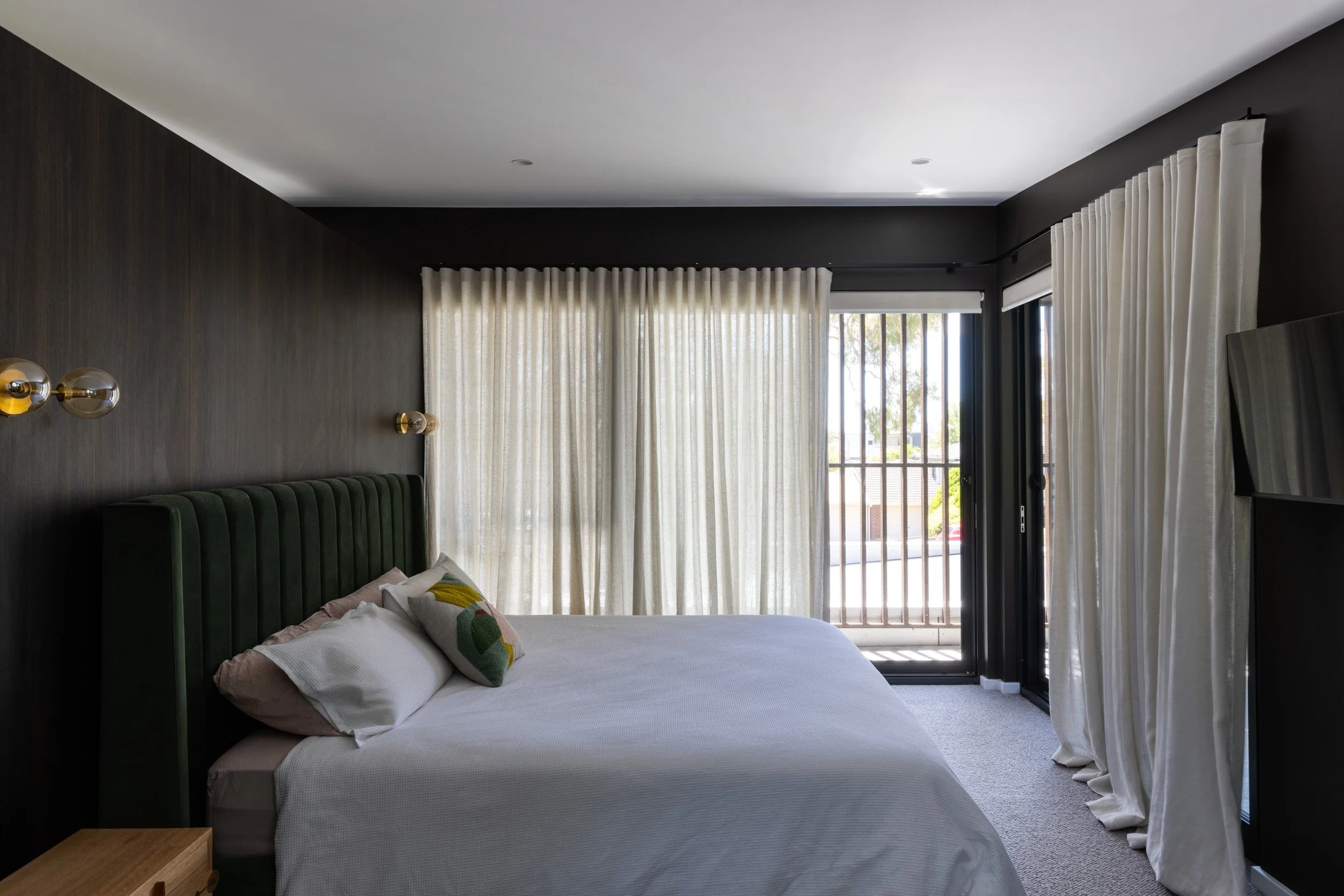 A modern bedroom with a bed, white bedding, and colorful pillows. The room has dark walls, a large window with cream curtains, and a sliding glass door leading to a balcony or outside area.