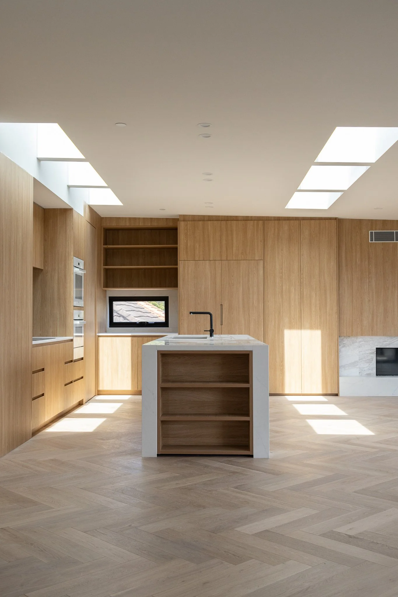 Modern kitchen with light wood cabinets, a white island with open shelves, black faucet, and a small window, illuminated by natural light and ceiling lights.