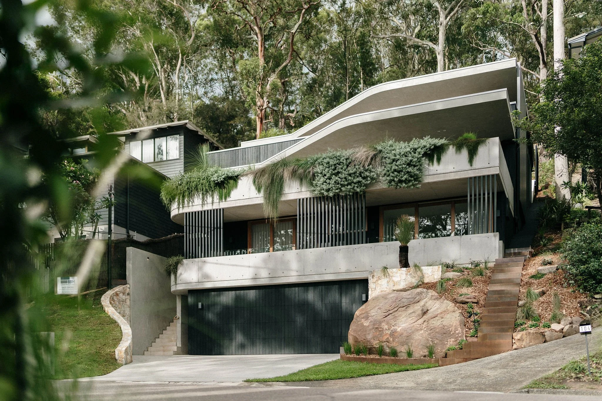 Modern multi-story house with curved architectural design, surrounded by trees and greenery, with a driveway and stairs leading up to the entrance.