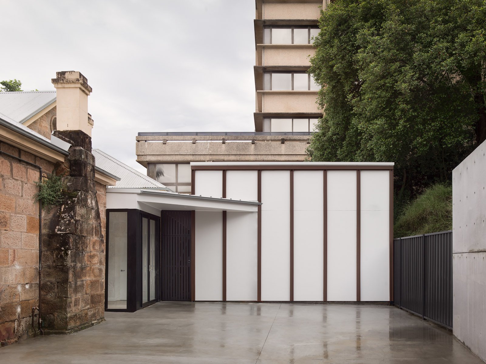 Modern building with white panels and brown framing, concrete patio, brick wall, and a large green tree against cloudy sky.