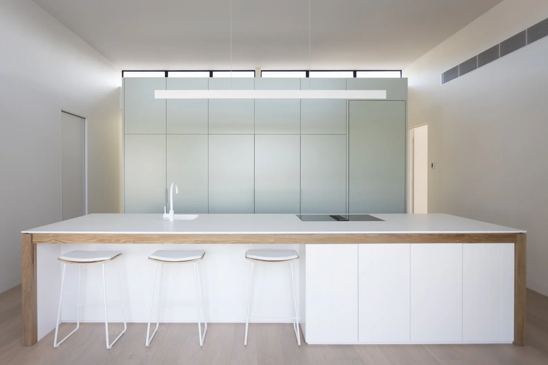 Modern minimalist kitchen with white island, three white stools, a white sink with a faucet, and a cooktop. Background features light-colored cabinets and a high windowed wall.