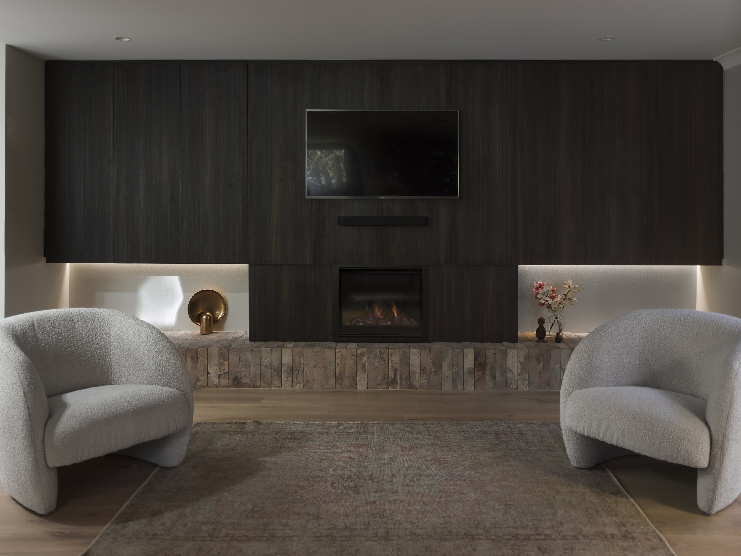 Living room with beige armchairs, a dark wood accent wall with a mounted flat-screen TV, a fireplace, and decorative vases and flowers.