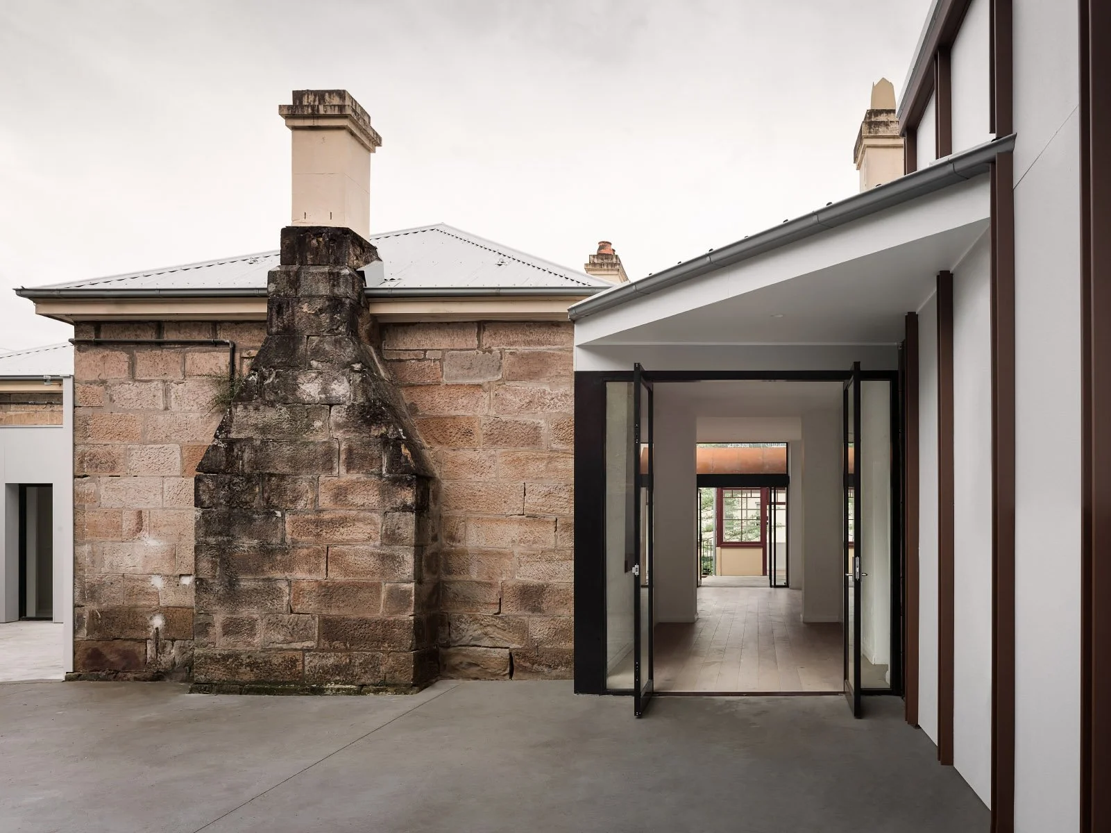 A modern building with a stone chimney and a contrast between old brick and contemporary white and brown exterior walls, with an open glass door leading inside.