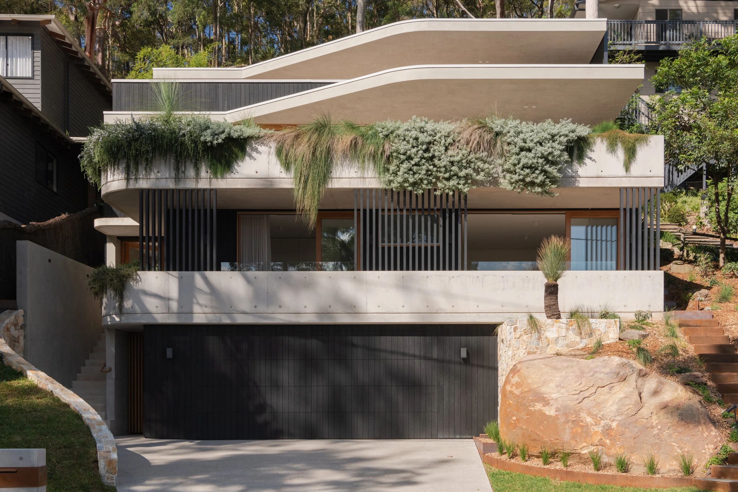Modern multi-story house with curved balconies, lush greenery, rocks, and a driveway in a wooded area.