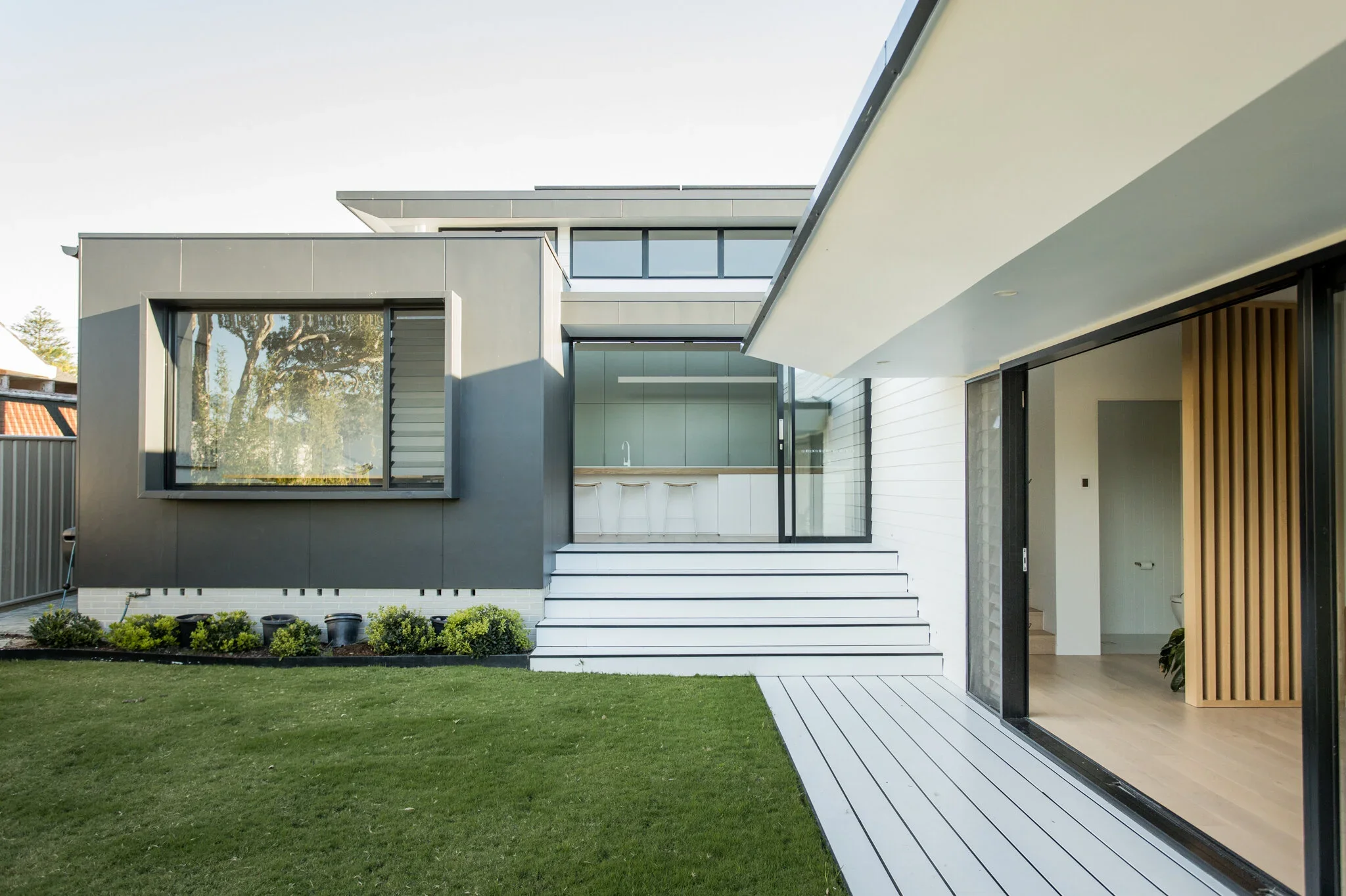 Modern house exterior with large sliding glass doors, a balcony, a staircase leading up to the house, and a lawn in the foreground