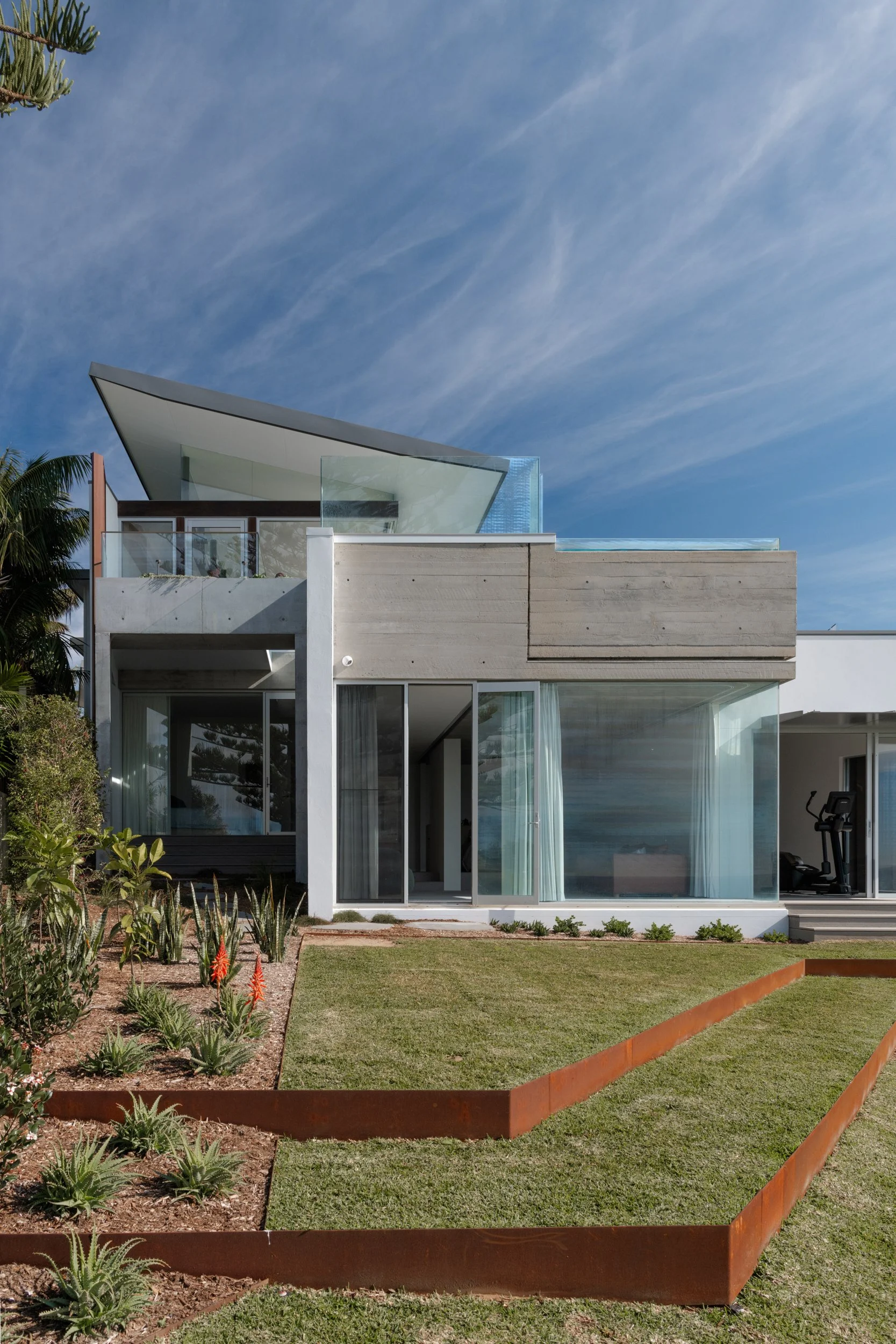Modern house with geometric roof design, glass walls, and a garden with plants and grass in the foreground.