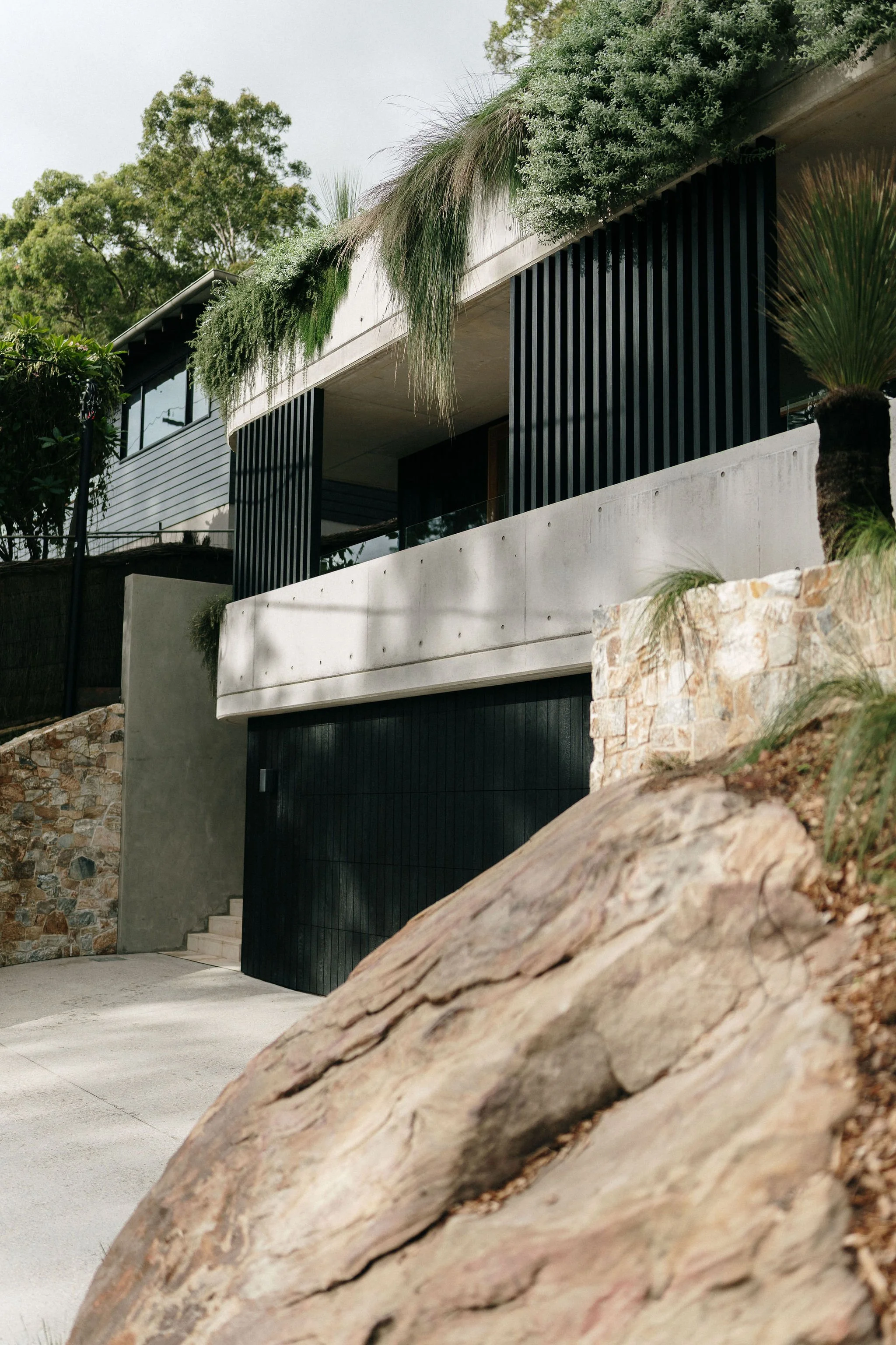 A modern house with black vertical slats and a concrete balcony, surrounded by green trees and plants, with a rocky foreground and an overcast sky.