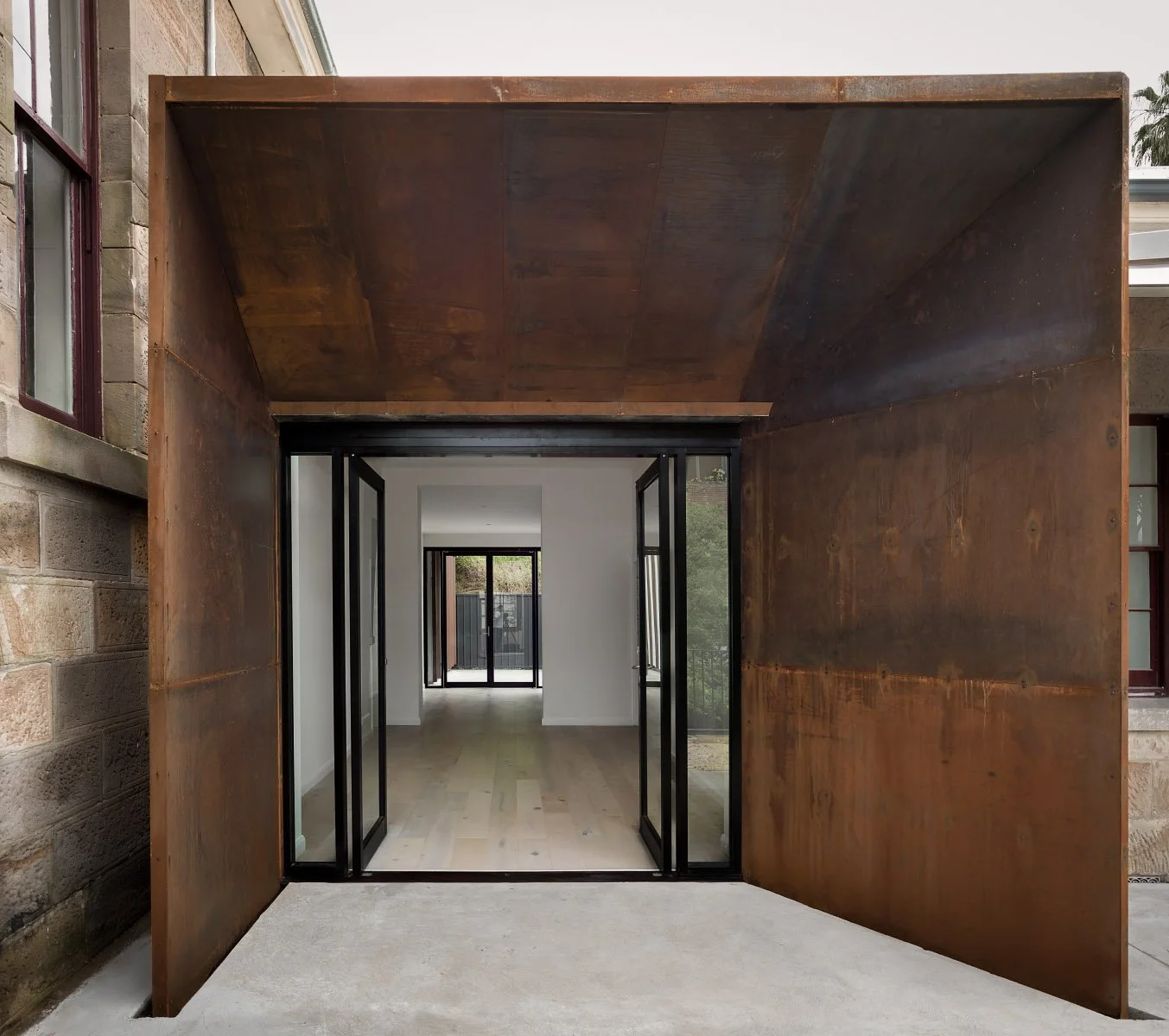 View of a modern building entrance with rust-colored metal walls and black-framed glass doors, leading to a white interior with large windows.