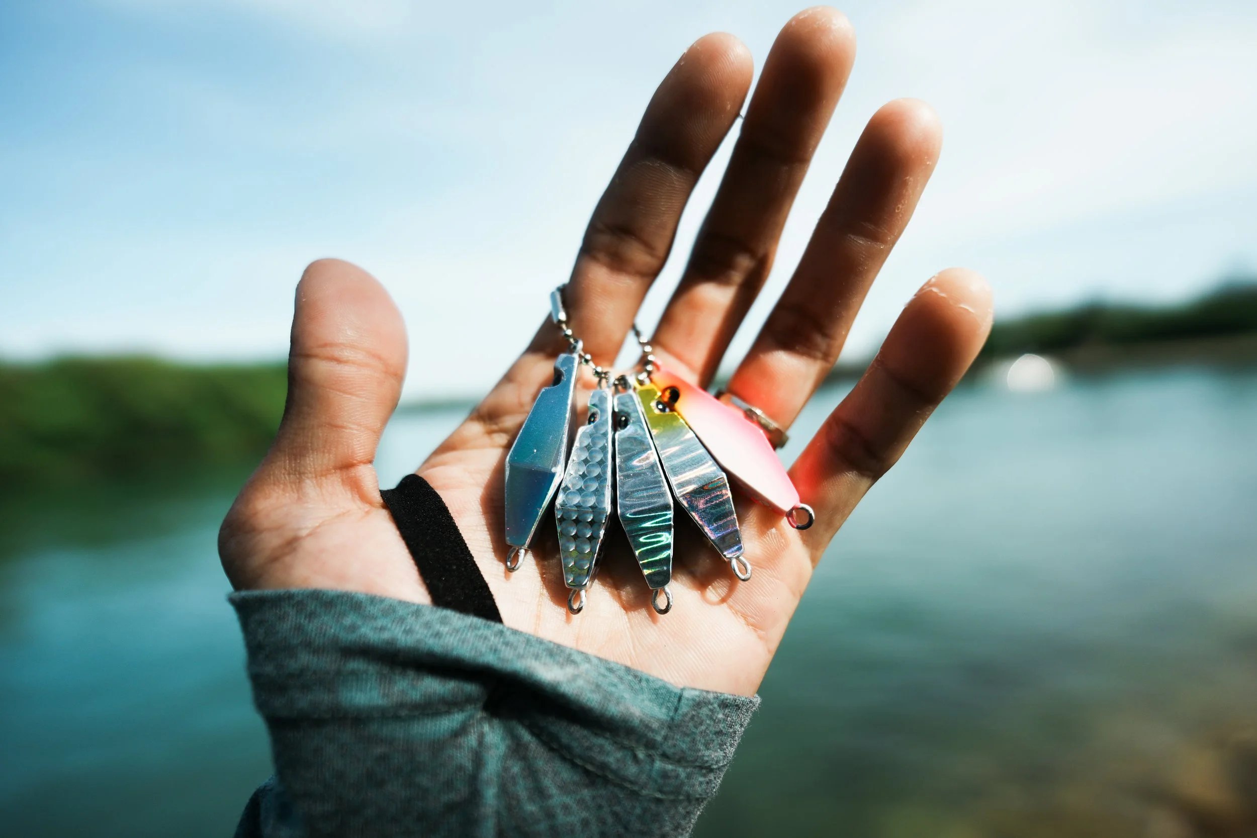 A hand holding a set of shiny fishing lures outdoors with a blurred natural background.