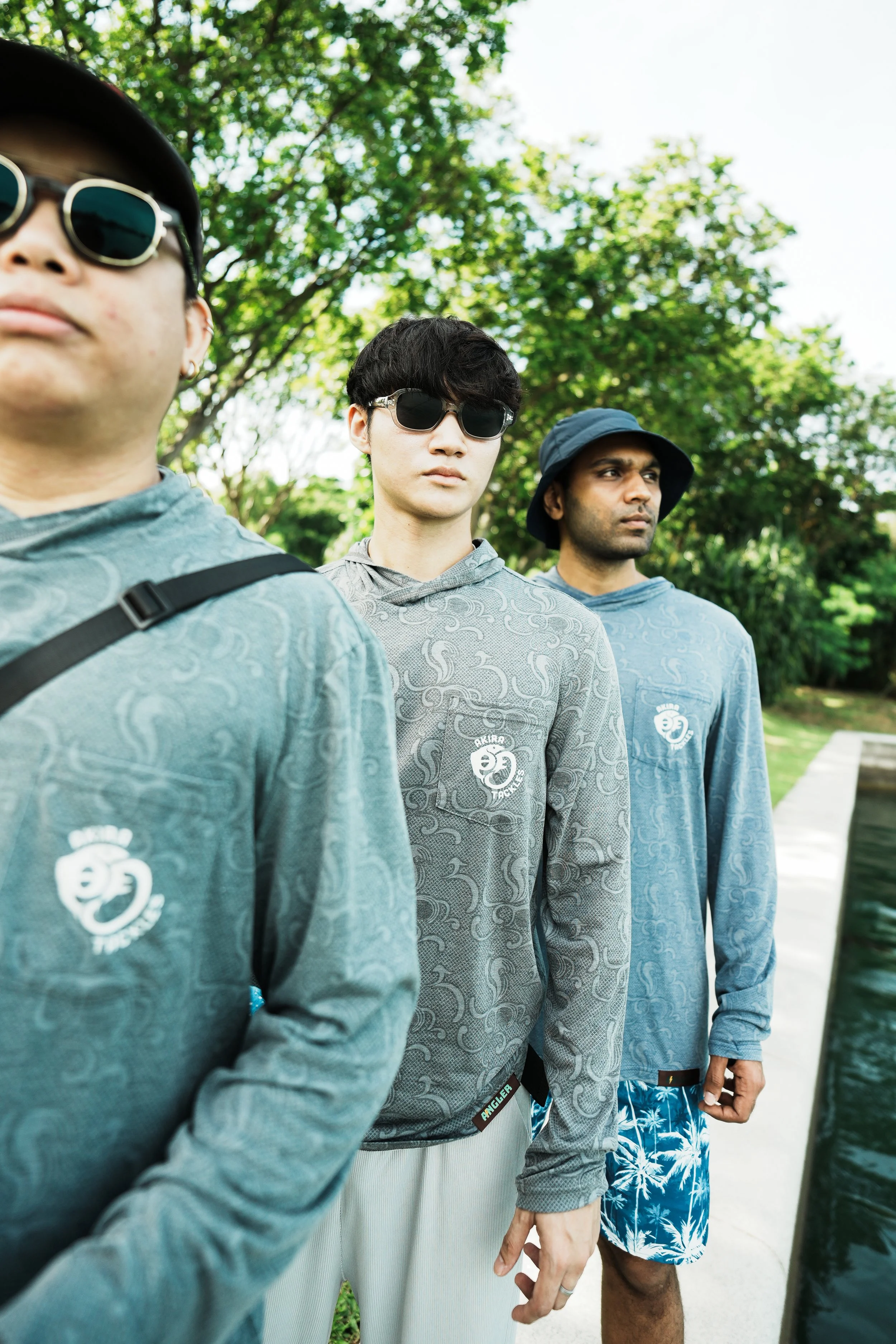 Three men standing outdoors on a sidewalk with green trees in the background. They are wearing casual clothes with matching shirts featuring a white logo that reads 'Akira Tackles,' and sunglasses.
