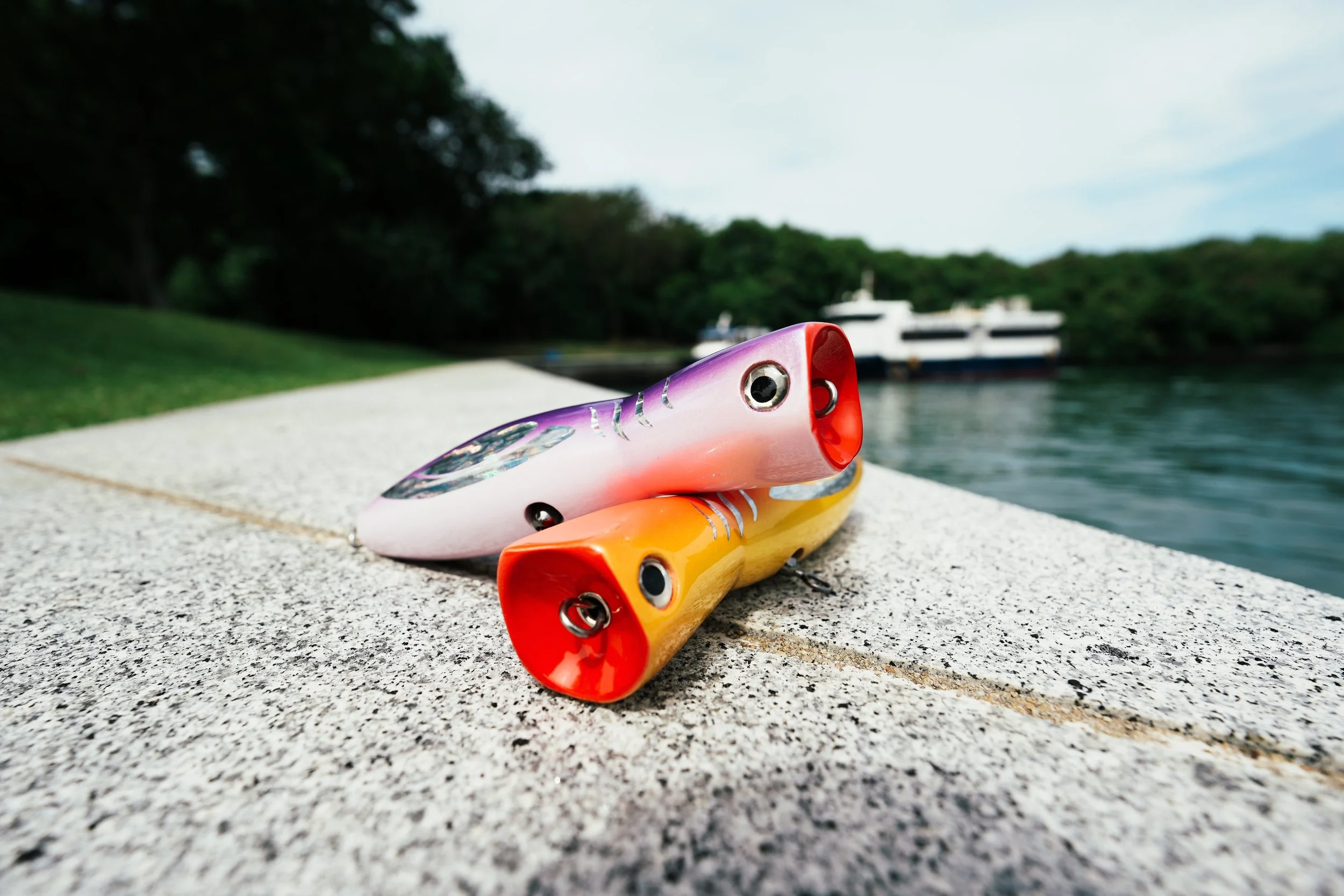 Colorful fishing poppers placed on a concrete ledge by a body of water with greenery and a boat in the background.