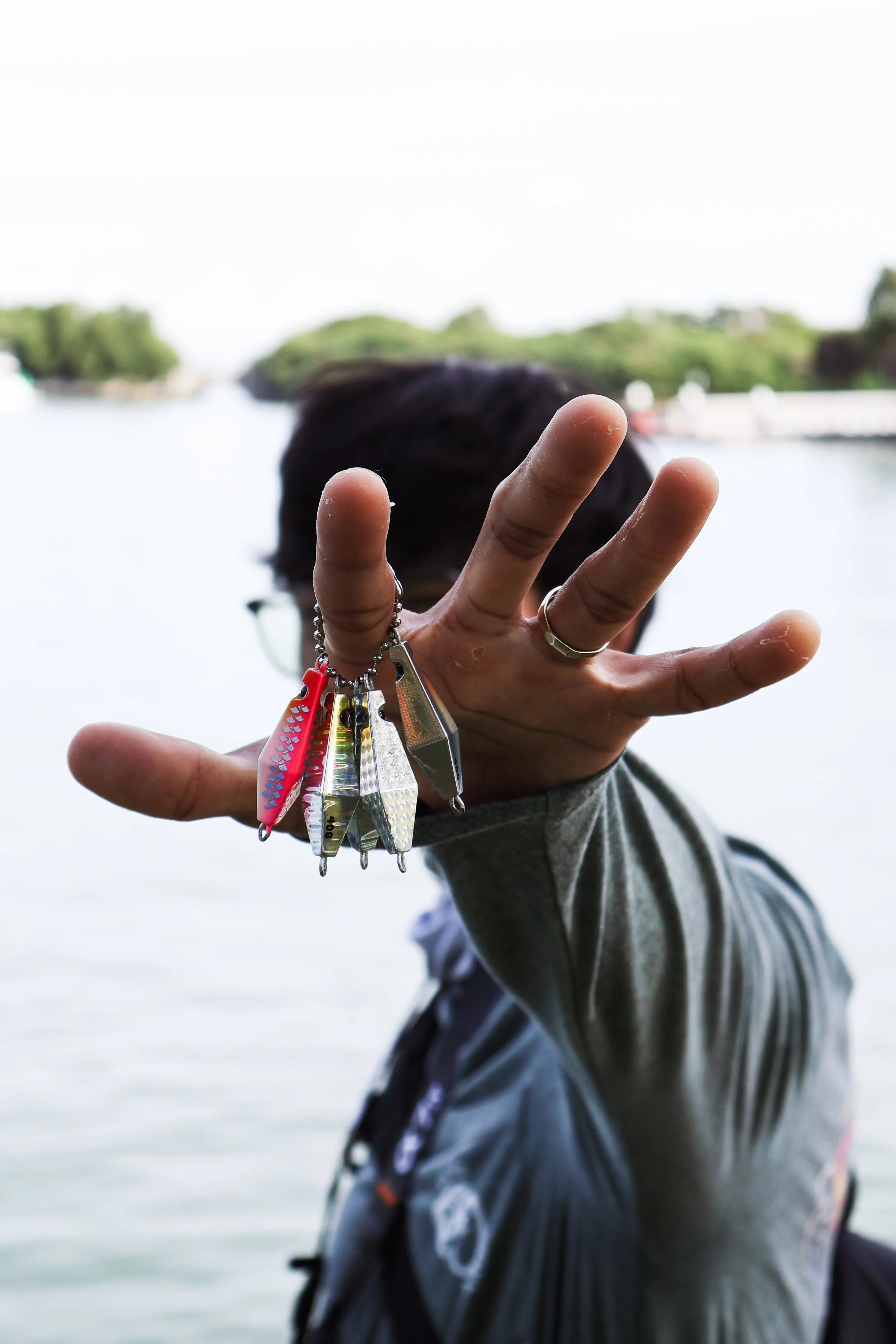 Person holding fishing lures in front of a body of water with a person wearing a life jacket visible in the background.