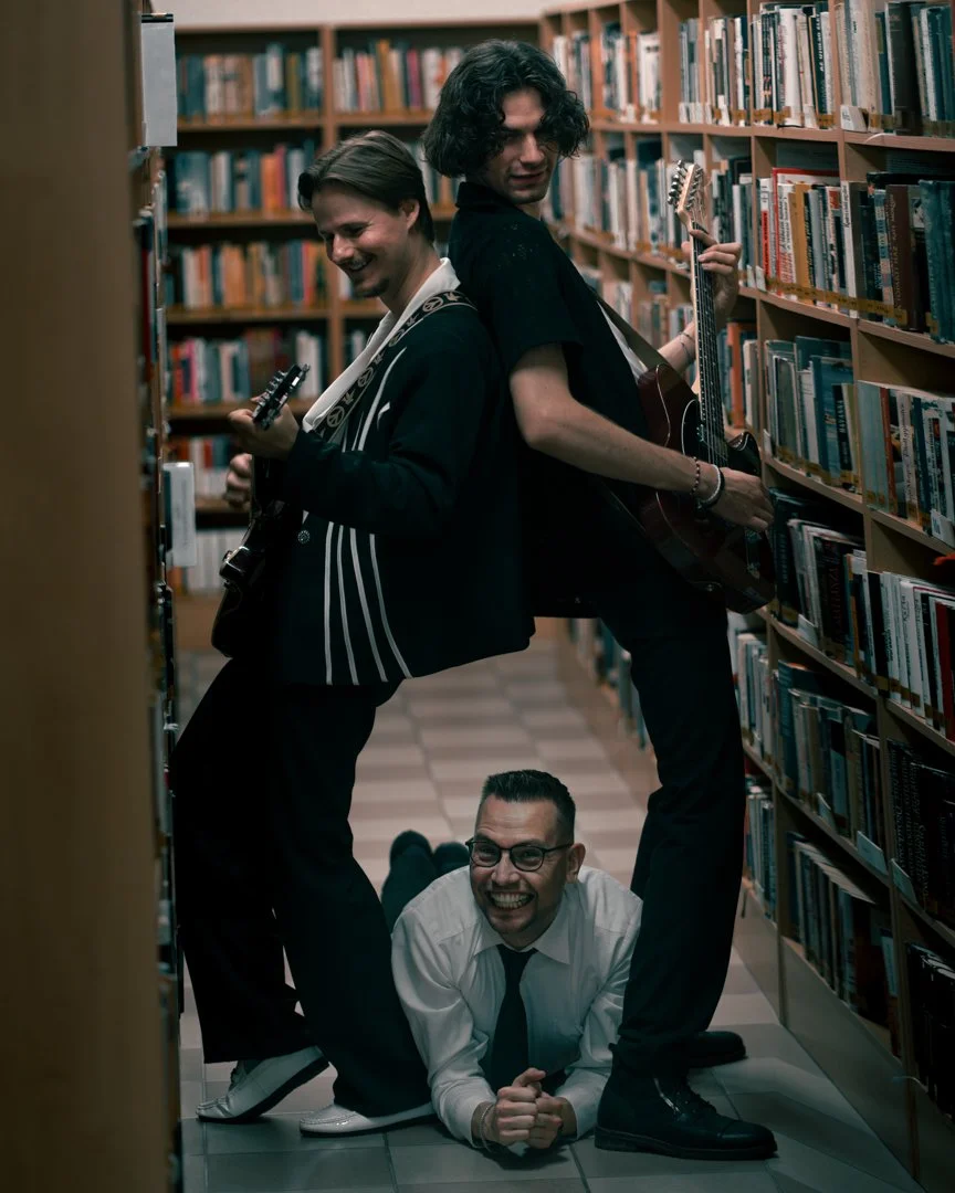 Three men with guitars in a library; two standing back-to-back with guitars, one lying on the floor under them, all smiling and appearing playful.