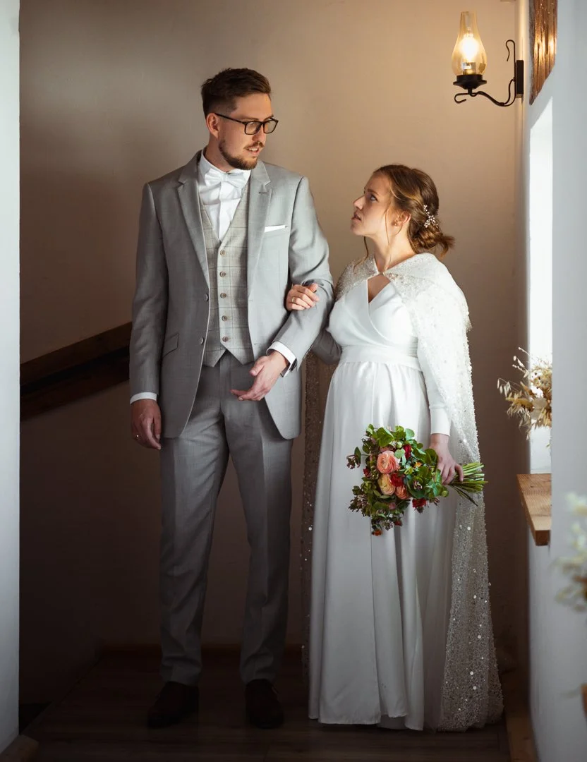 A bride and groom standing indoors, holding each other's arms, with the bride holding a bouquet of flowers and looking at each other.