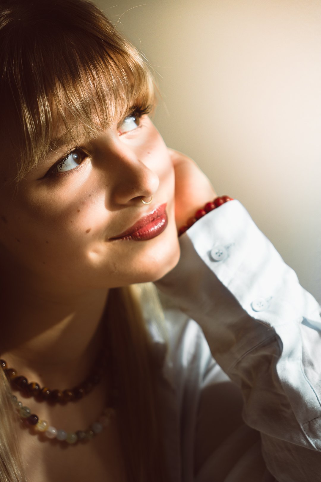 A young woman with red hair and fair skin, wearing a white shirt, red beaded bracelet, and multicolored beaded necklace, gazing thoughtfully to the side with natural light illuminating her face.