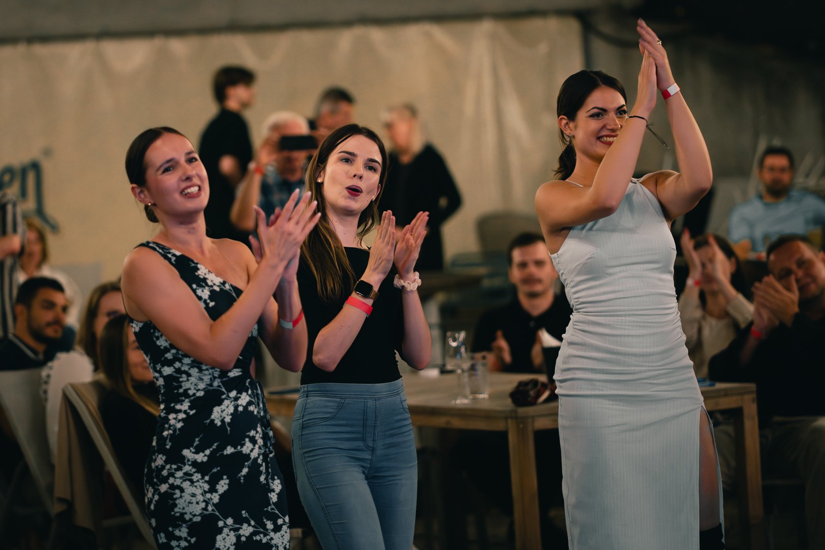 Three women standing and clapping at an indoor event with an audience in the background.