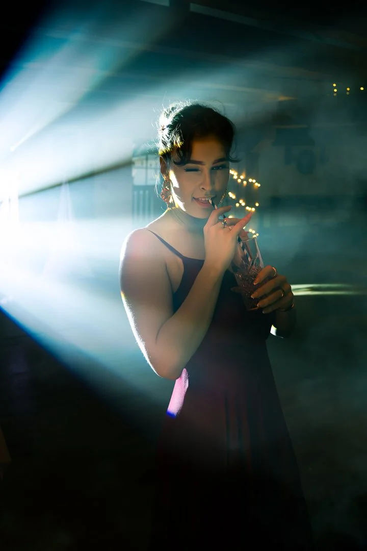 A woman winks and sips from a glass in a dimly lit space with colorful stage lights and festive string lights in the background.