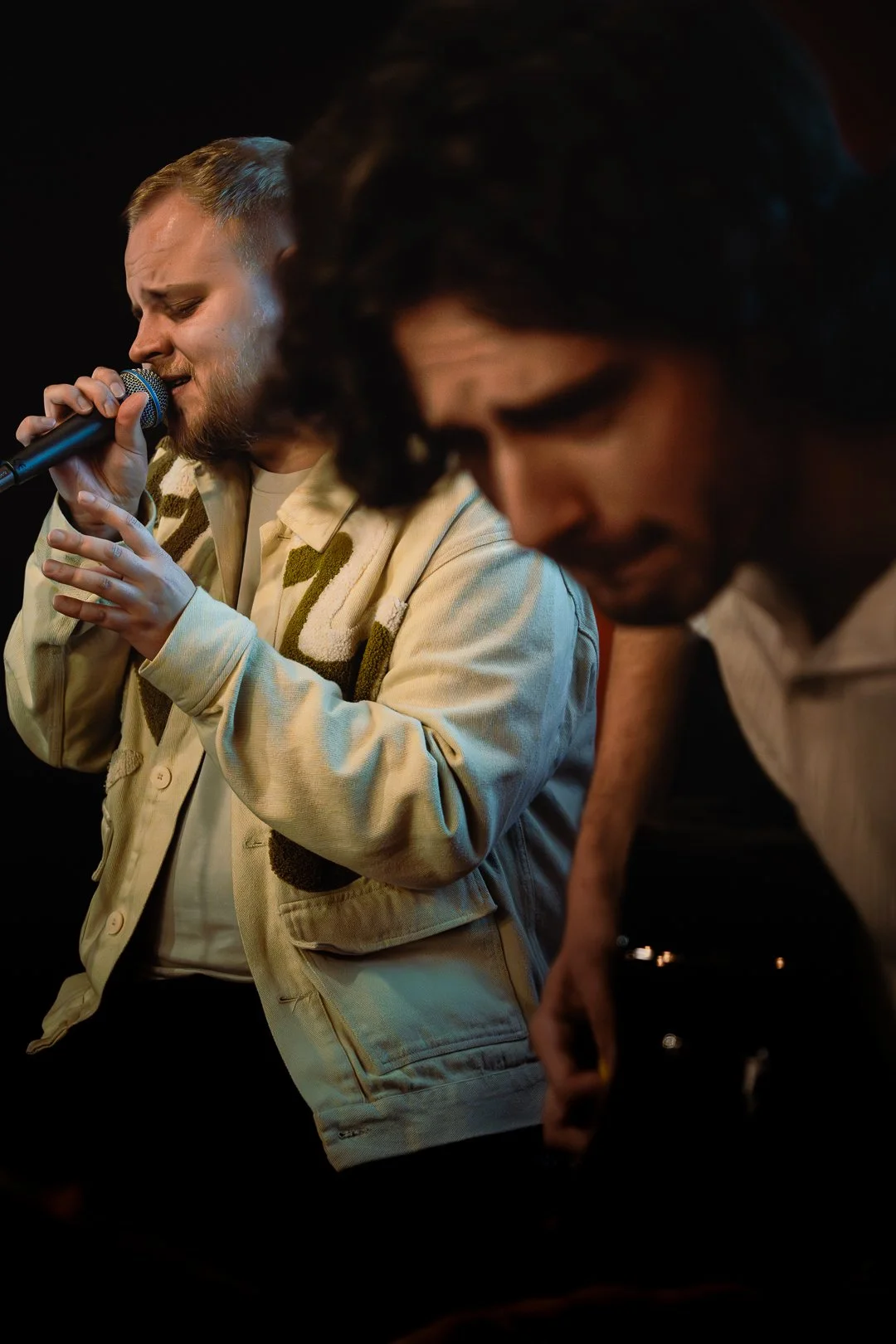 Two men singing passionately, one with a microphone, in a dimly lit setting.