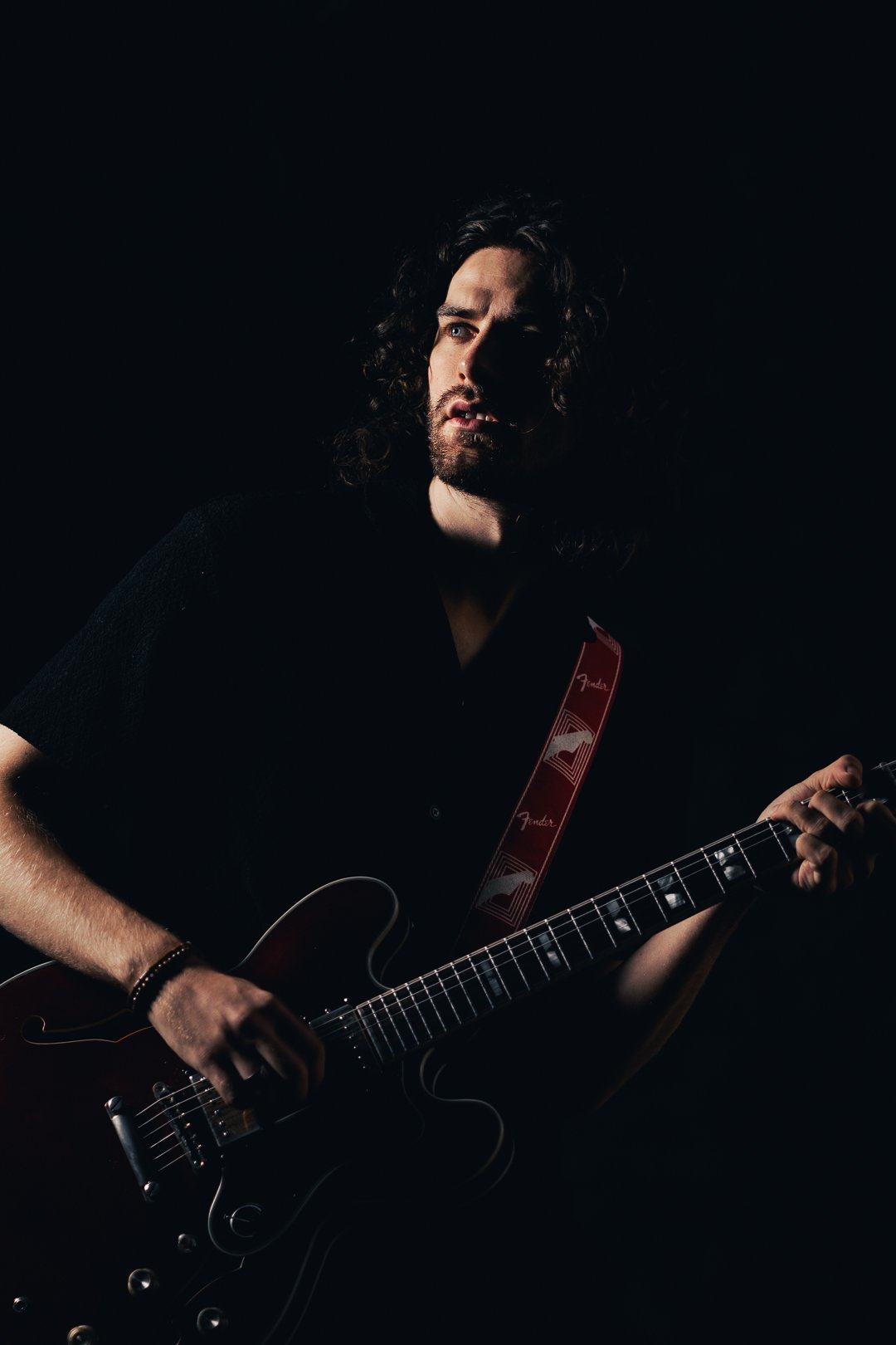 A man with long curly hair and a beard playing an electric guitar in a dark setting, with dramatic lighting highlighting his face and upper body.