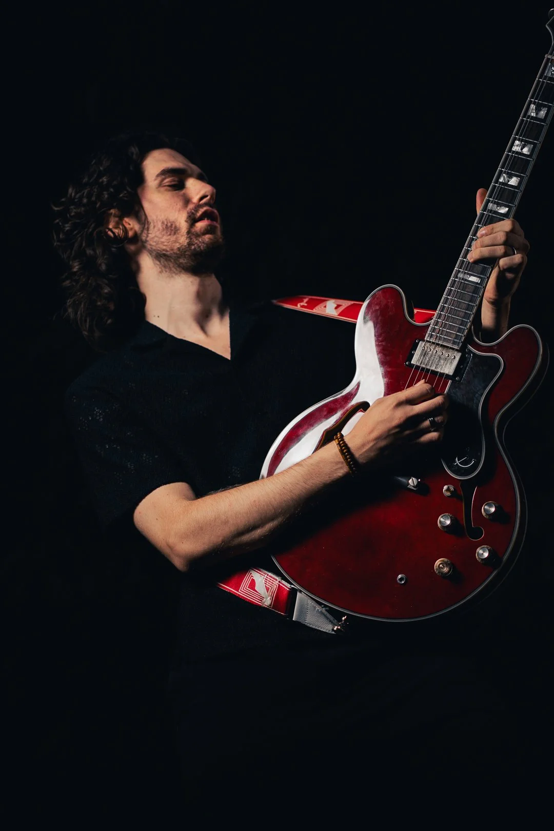 A man with long curly hair and a beard playing an electric guitar against a black background.