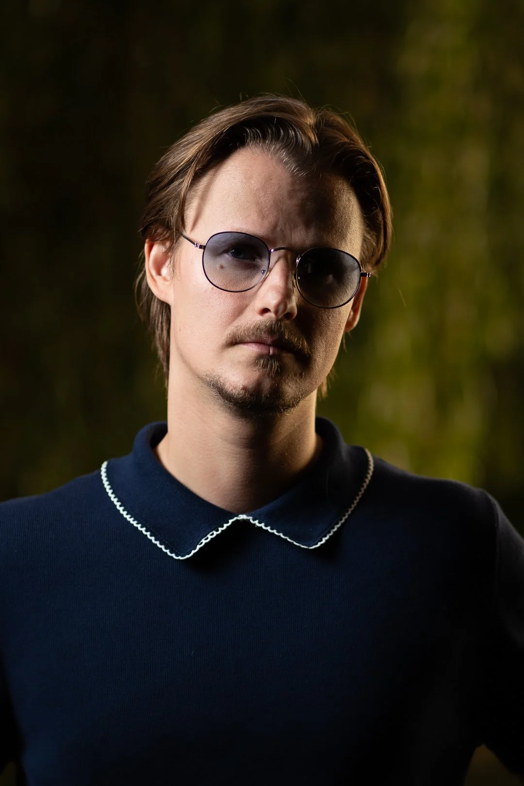 A young man with brown hair, wearing sunglasses and a navy blue top with a white-trimmed collar, standing outdoors at dusk or night with blurred greenery background.