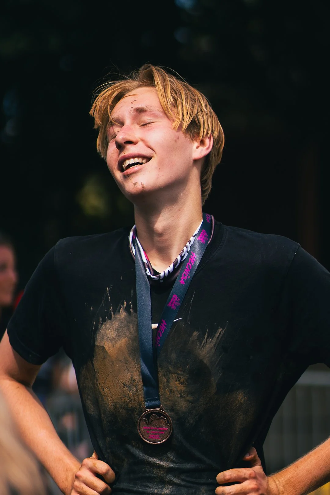 A young male athlete with a medal around his neck, smiling with his eyes closed after a race, wearing a black T-shirt with mud stains.