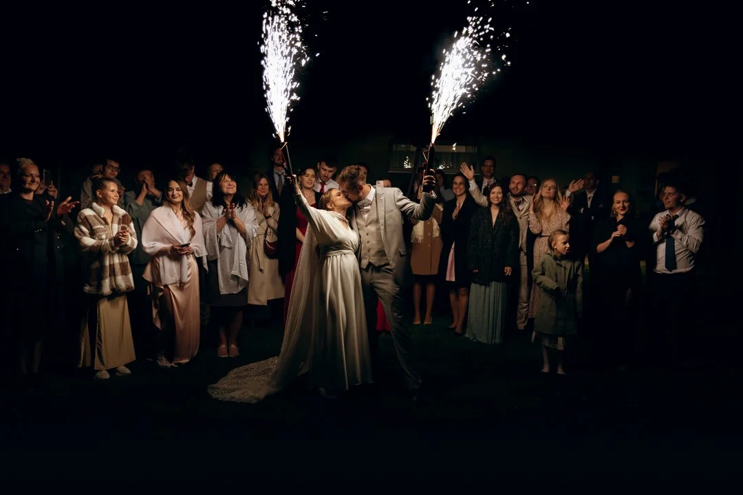 A bride and groom share a kiss while holding sparklers at their wedding reception, surrounded by guests clapping and celebrating at night.