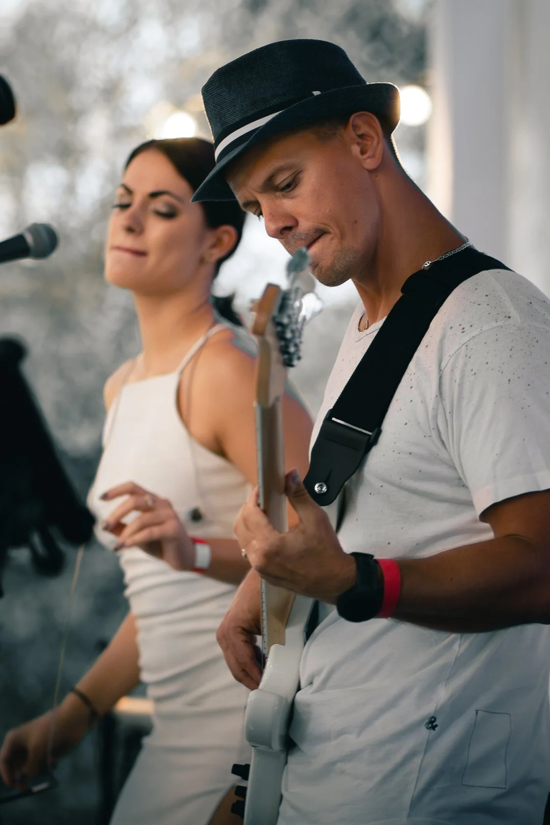 A man wearing a black hat and a white shirt playing an electric guitar, with a woman in a white dress singing into a microphone in the background.