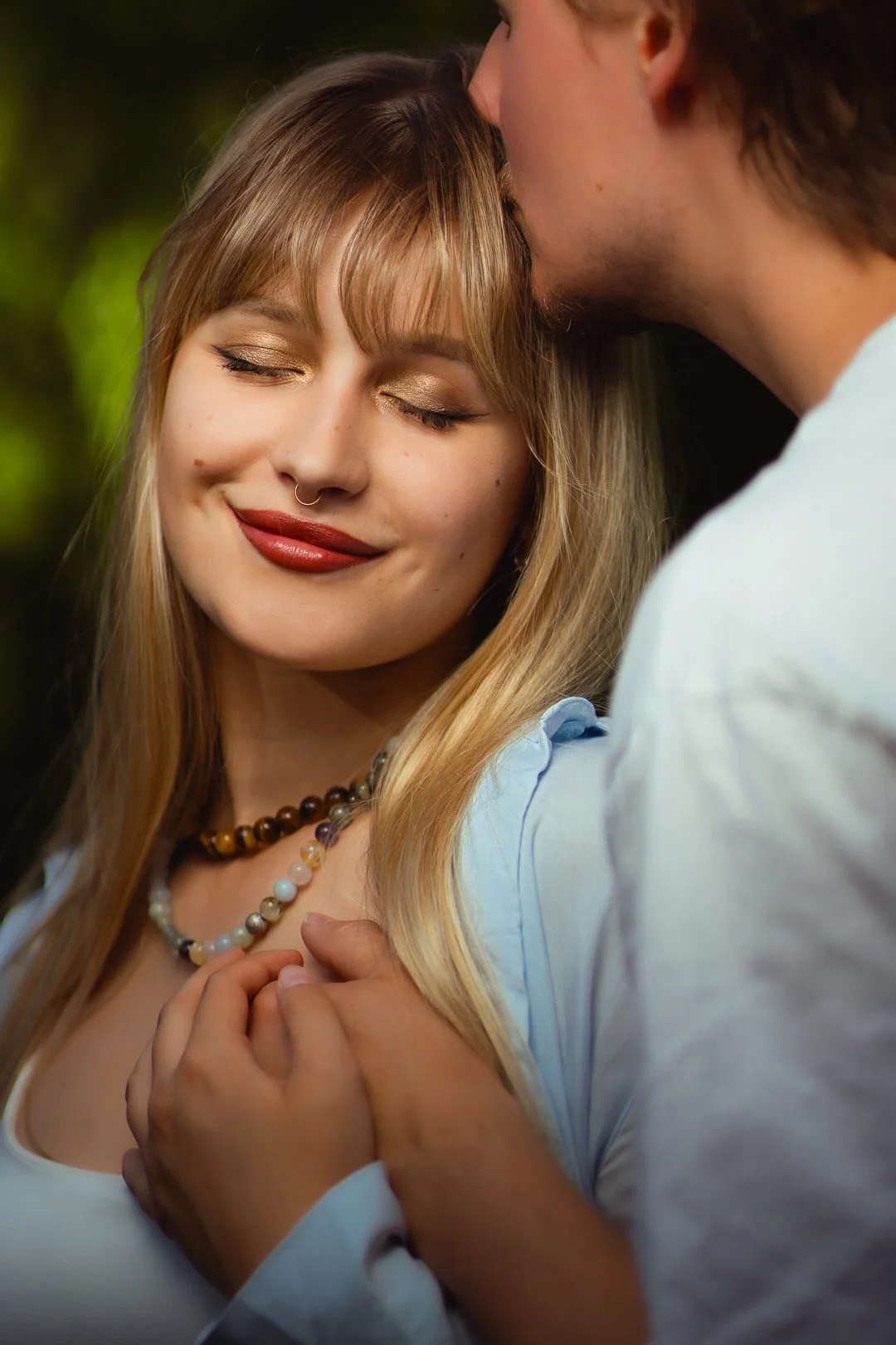 A woman with long blonde hair, closed eyes, smiling, wearing layered beaded necklaces, being kissed on the forehead by a man with short hair, both wearing white shirts, holding hands, with a soft, blurred green background.