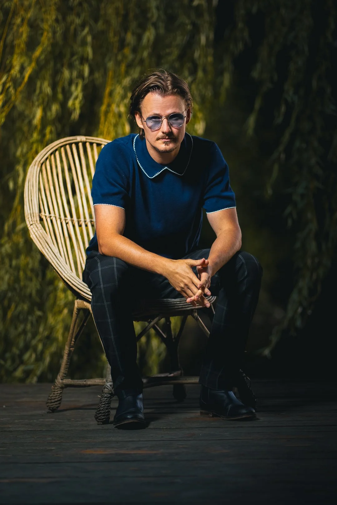 A young man with glasses and dark hair sitting on a wicker chair outdoors, wearing a blue shirt and black plaid pants.