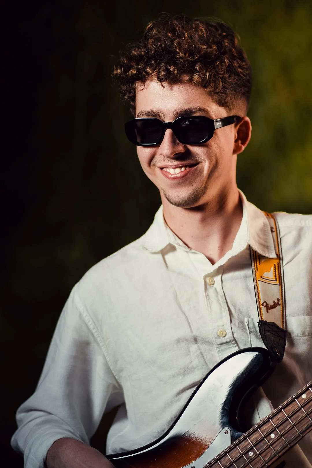 A young man with curly brown hair wearing black sunglasses and a white shirt, smiling and holding a Fender electric guitar.