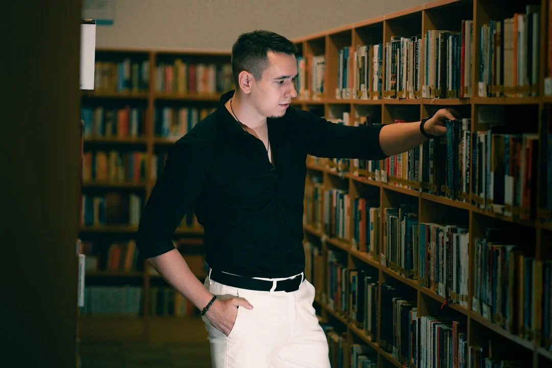 A young man in a black shirt and white pants browsing books in a library, looking at a book on a shelf.