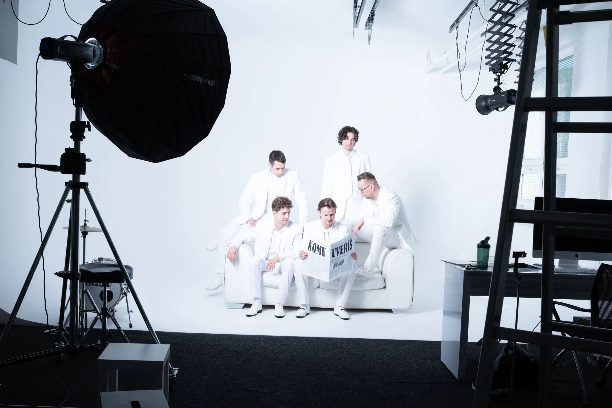 A group of six men dressed in white suits sitting and standing on and around a white couch in a photography studio. One man is reading a newspaper while the others are looking at him or each other. Studio equipment, including lights, a ladder, and a table with bottled water, is visible.