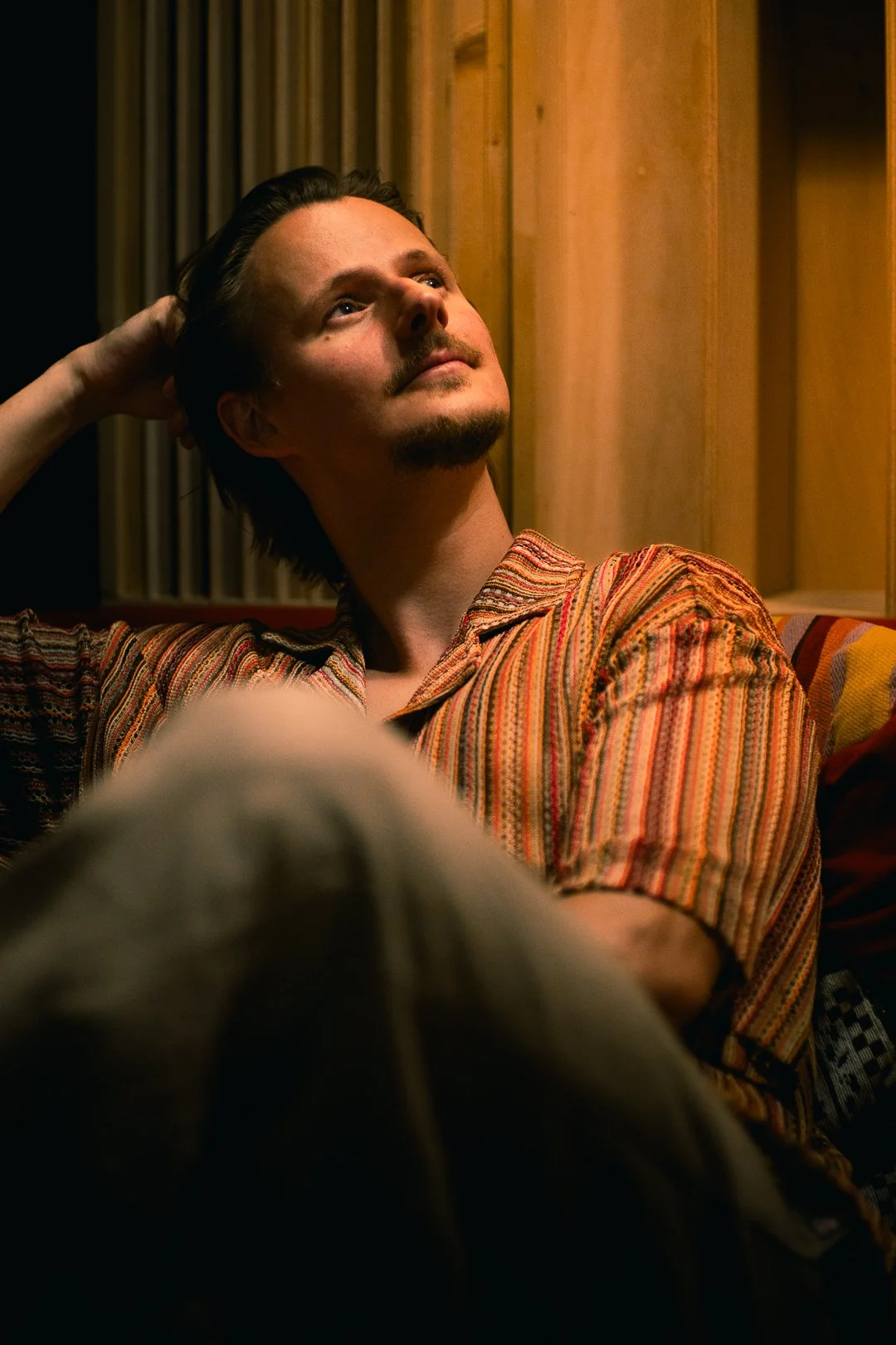 A man with dark hair, mustache, and goatee, wearing a colorful striped shirt, sitting indoors against wooden paneling, looking upwards thoughtfully.