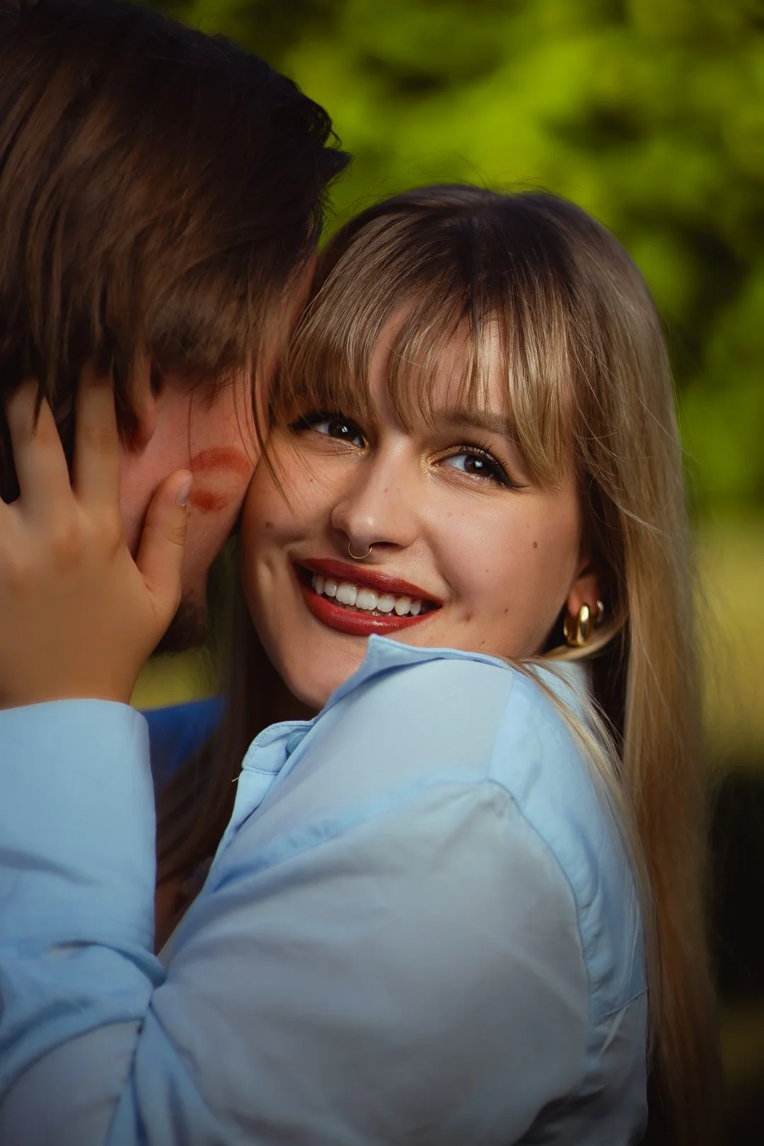 A woman with blonde hair, a nose ring, and gold earrings smiling while whispering into a man's ear, who has brown hair and is not fully visible.