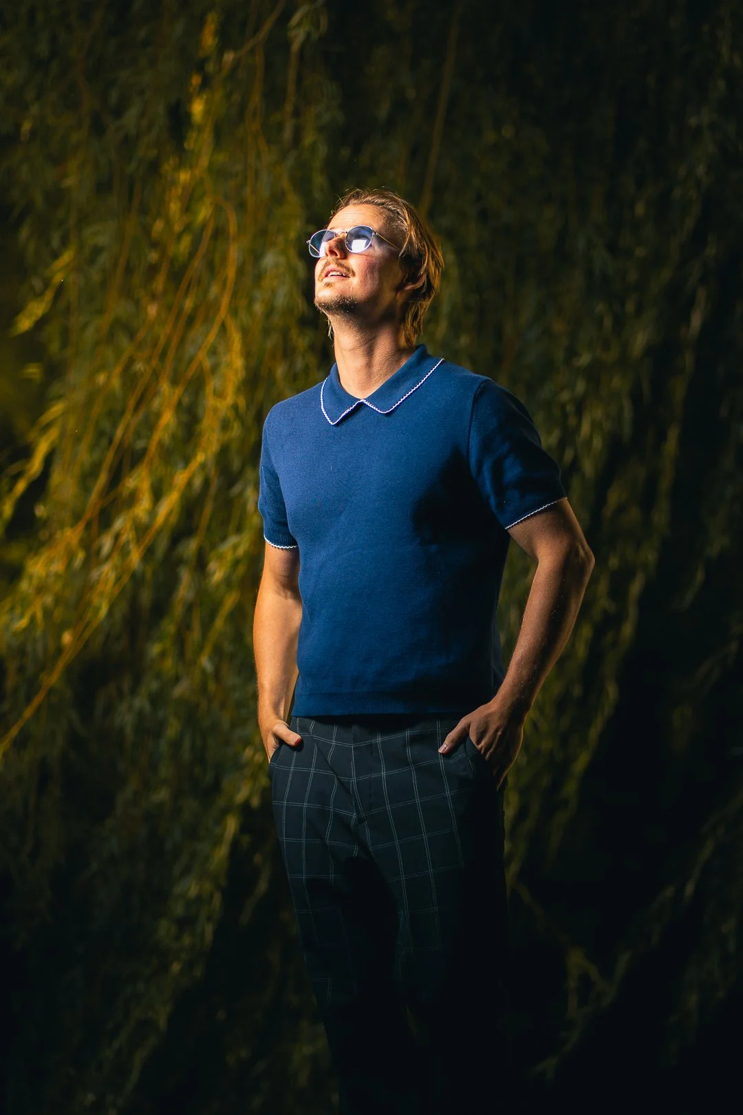 A man in a blue collared shirt and plaid pants standing outdoors at night with his hands in his pockets, looking upward with sunglasses on, in front of a dark, natural background.