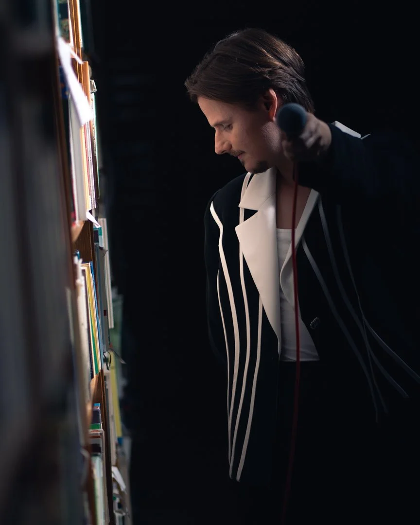 Man in a black and white striped blazer looking at books on a library shelf while holding a microphone.