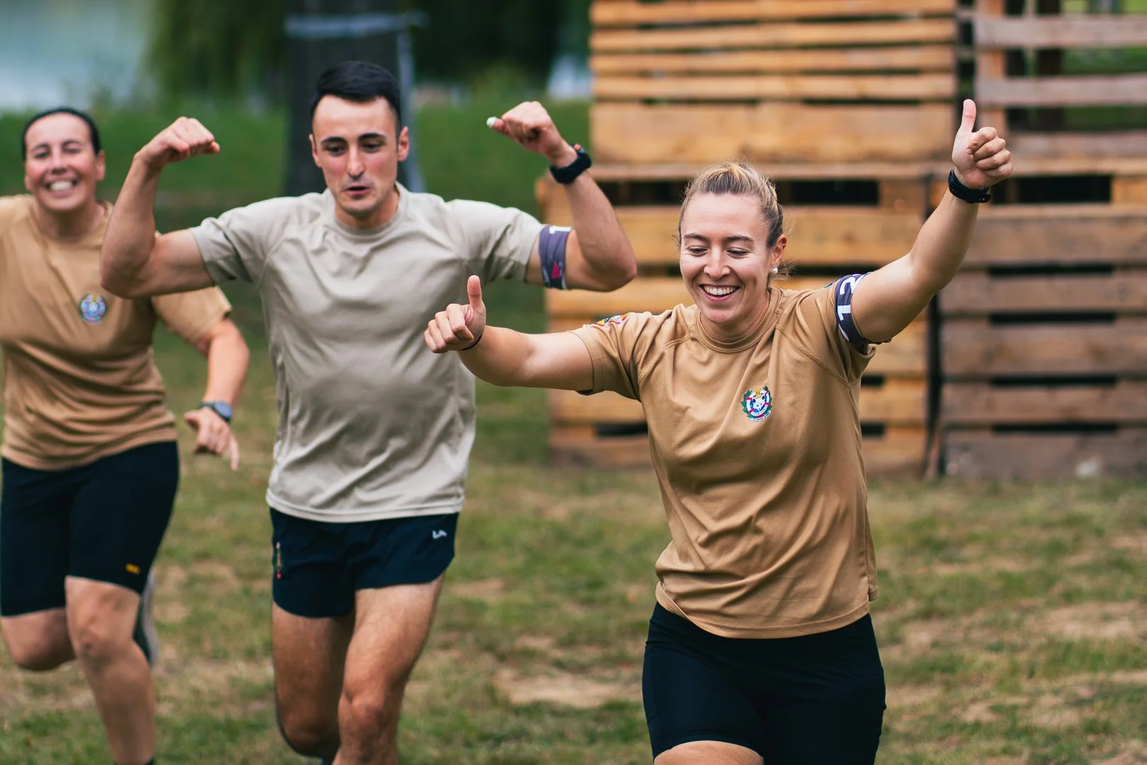 Three people running outdoors on grass, smiling and celebrating, with a woman in the foreground giving a thumbs up.