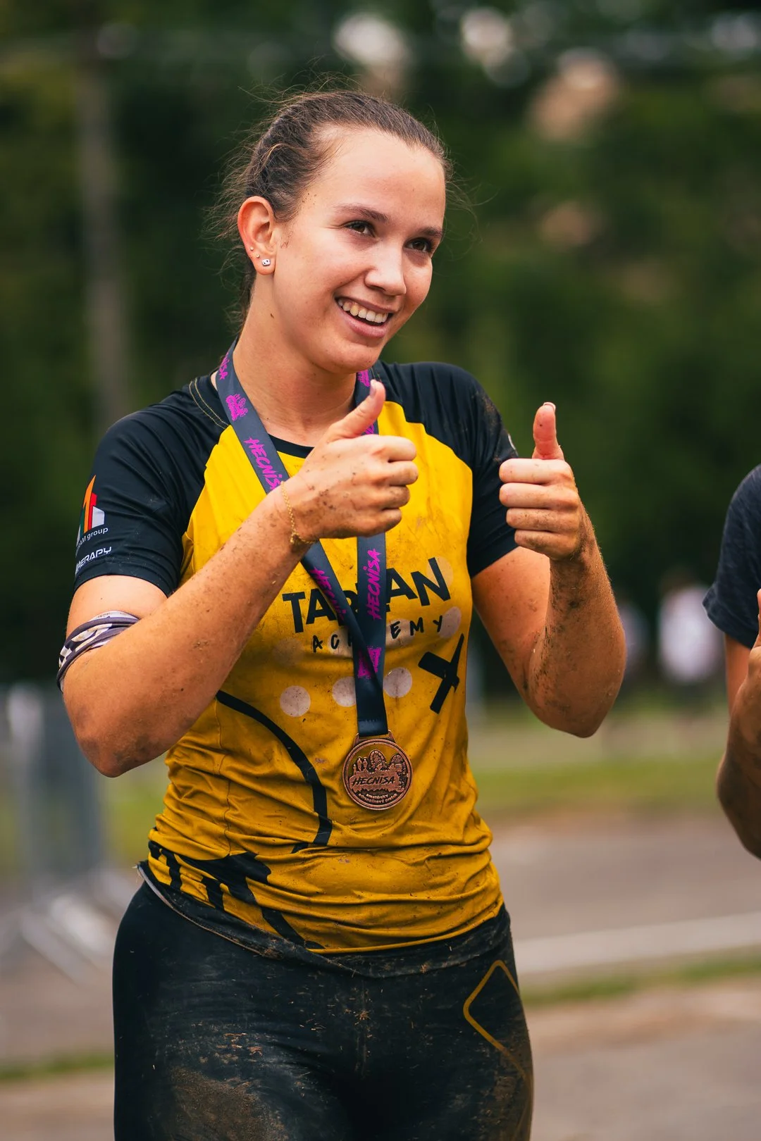 A young woman smiling and giving thumbs up, wearing a yellow and black sports shirt with a medal around her neck during an outdoor event.