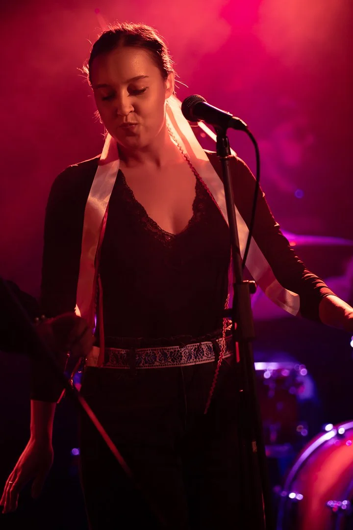 A woman singing into a microphone on stage with pink and purple lighting.