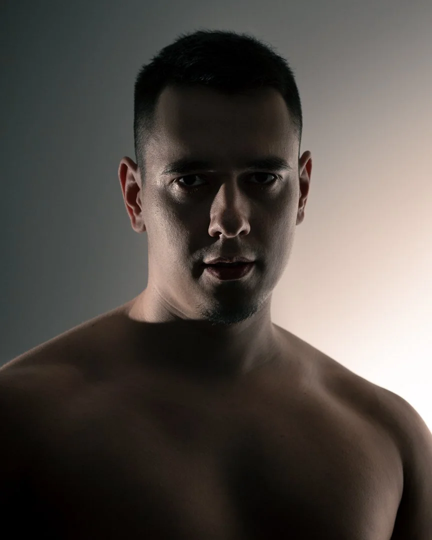 A shirtless man with short black hair looking at the camera with a serious expression, lit from behind creating a shadowed front.
