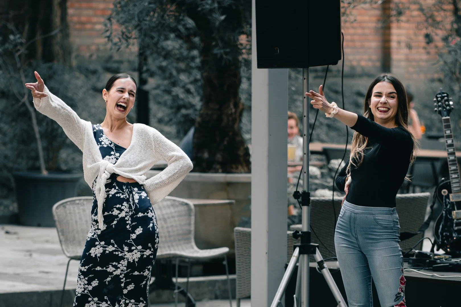 Two women smiling and dancing outdoors near a speaker and a guitar, with other people seated at tables in the background.
