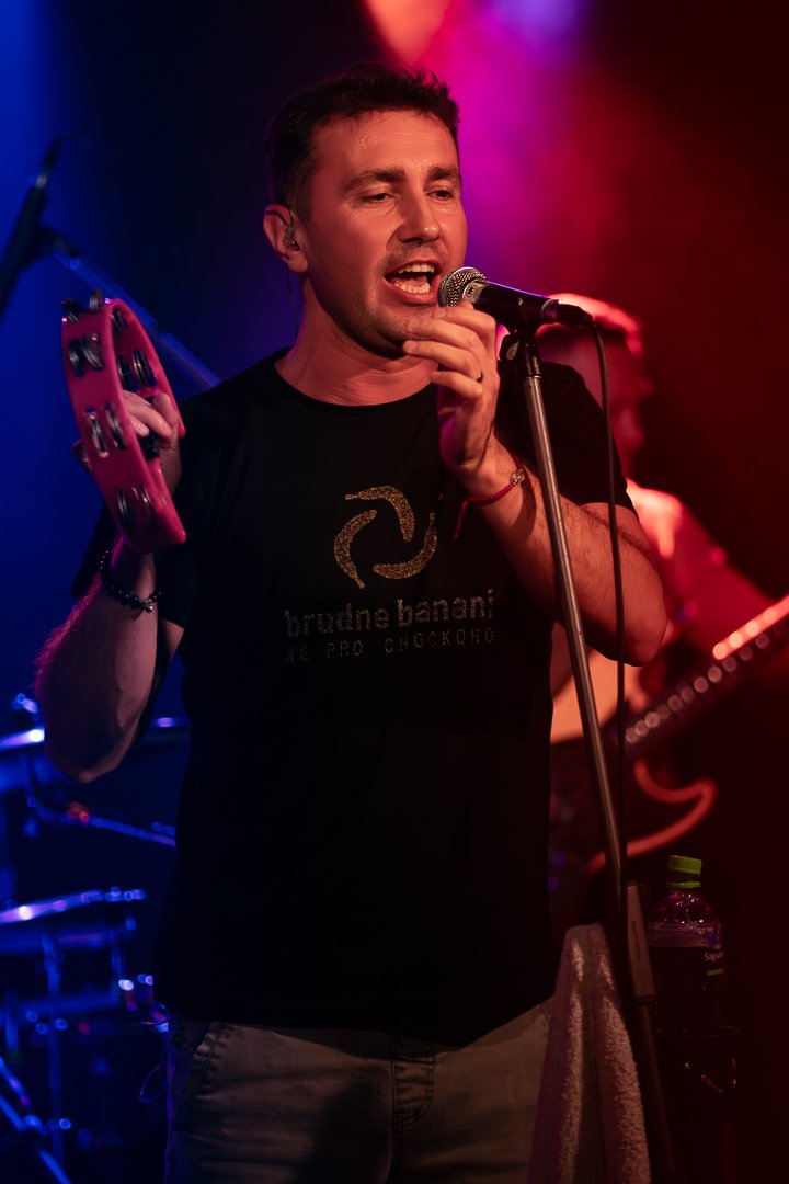 A man singing into a microphone while holding a tambourine in a dimly lit concert setting with red and purple stage lighting. He is wearing a black t-shirt with a logo.
