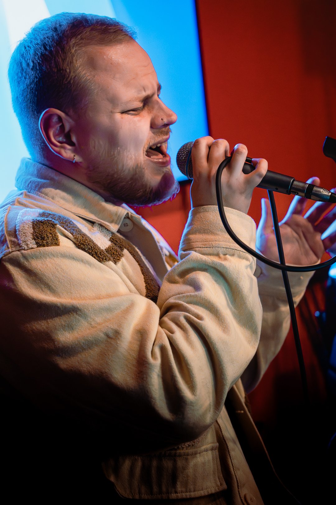 A man with short hair, a nose piercing, and earrings is passionately singing into a microphone, with his eyes closed and a serious expression, against a colorful background.