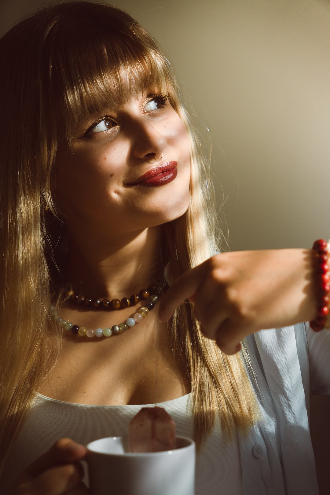 A woman with long blonde hair, wearing necklaces and a white top, looks up thoughtfully while holding a red beaded bracelet in her right hand and a white mug with a dessert inside in her left hand.