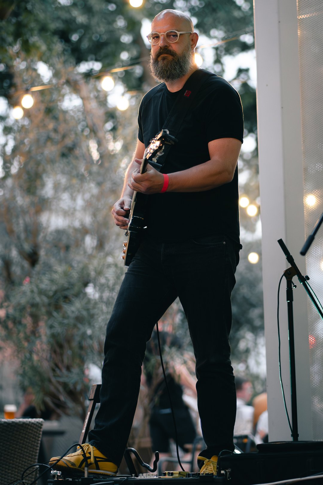 Man with a gray beard and glasses playing an electric guitar during an outdoor event at dusk, with blurred trees and string lights in the background.