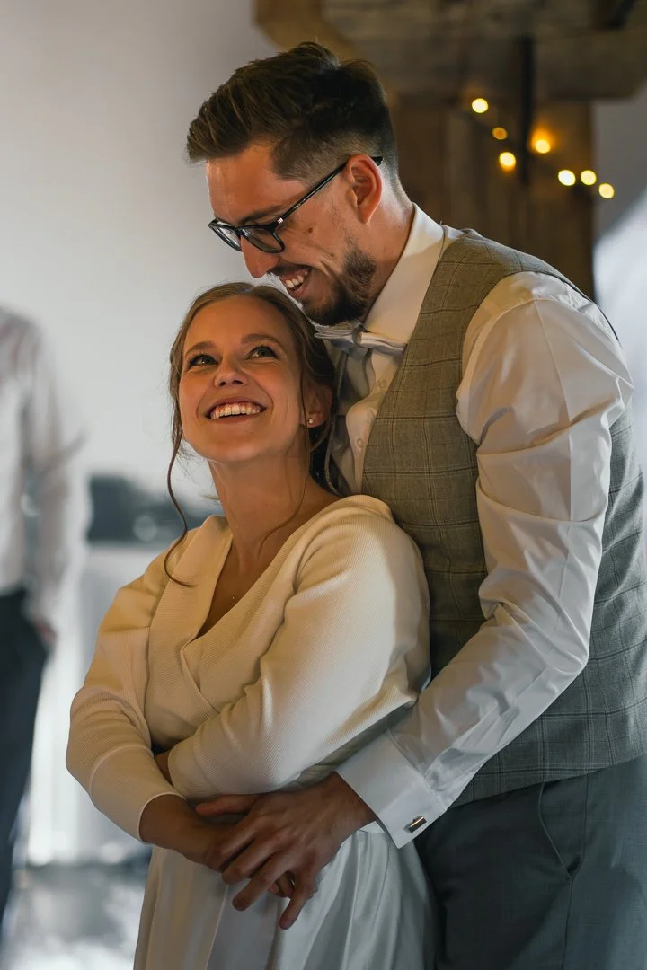 A man and woman share a joyful moment, with the man hugging the woman from behind. They are smiling brightly, looking at each other, in a warmly lit indoor setting with a rustic wooden ceiling and some decorative lights in the background.