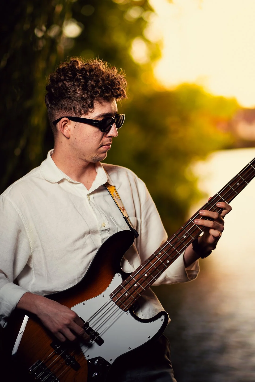 A young man with curly hair and sunglasses is playing an electric bass guitar outdoors during sunset, with trees and water in the background.