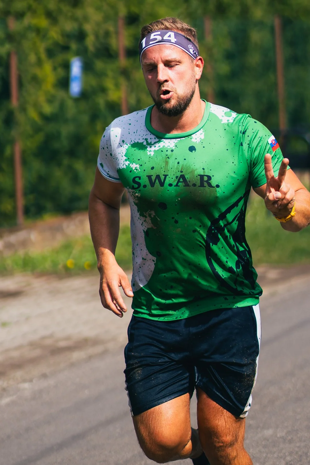 A man running outdoors in a green and white shirt with 'S.W.A.R.' printed on it, and making a peace sign with his right hand, wearing a headband and shorts.